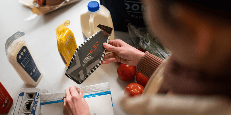 A person holds an AeroFlexx Pak over a table with groceries including a milk bottle and tomatoes.